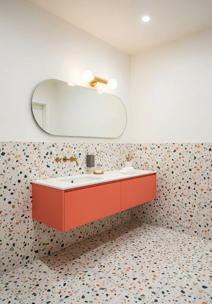 A playful and contemporary restroom design celebrating the beauty of terrazzo. Large-scale terrazzo with colorful aggregate covers the floor and extends halfway up the walls in a wainscoting effect. A simple, floating vanity in a complementary solid color allows the terrazzo to be the hero. Fixtures are modern and minimalist in a brushed brass finish. A large, pill-shaped mirror adds a soft, modern touch. A bright, evenly lit, wide-angle shot showcases the vibrant pattern and cheerful character of this material-driven design.