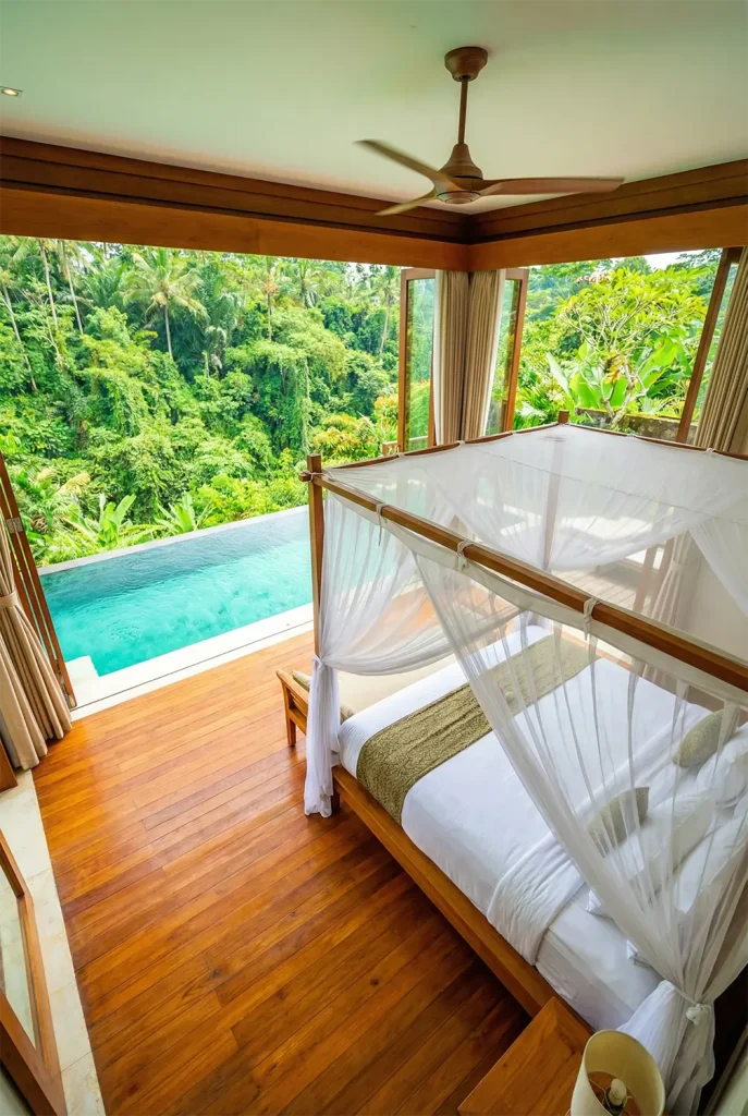 A breathtaking wide-angle perspective of an open-air tropical bedroom in Bali. The room has no walls on two sides, looking directly out onto a lush jungle and a private infinity pool. A canopy bed with mosquito netting sits on a polished teak floor. A ceiling fan spins lazily above. The lighting is bright and natural, capturing the vibrant greens of the rainforest and the rich browns of the wood.